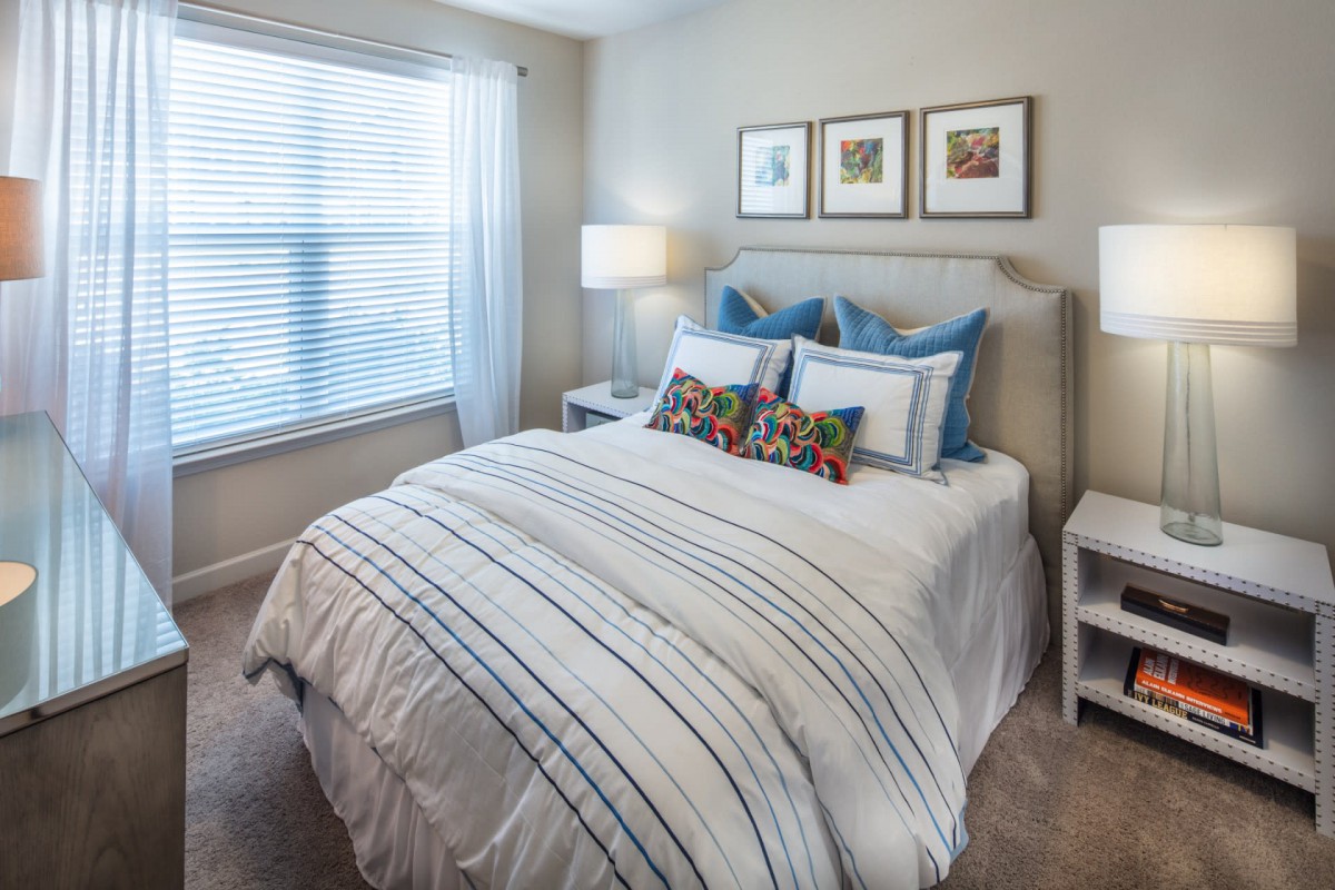 A cozy and well-decorated bedroom with a neatly made bed, featuring striped bedding and colorful pillows. The room is brightened by natural light from a large window and complemented by framed artwork above the headboard.