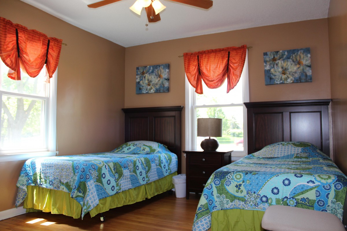 A cozy bedroom with twin beds featuring colorful bedding and a central nightstand under an orange-curtained window.