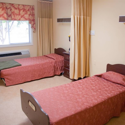 A hospital room featuring two single beds with red coverings and a window adorned with curtains.