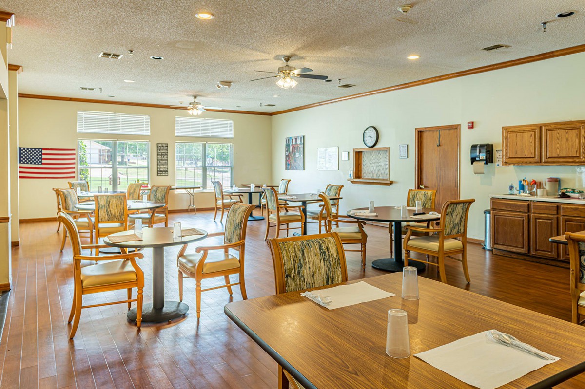 An empty dining room with several tables and chairs, featuring large windows and a wooden floor.