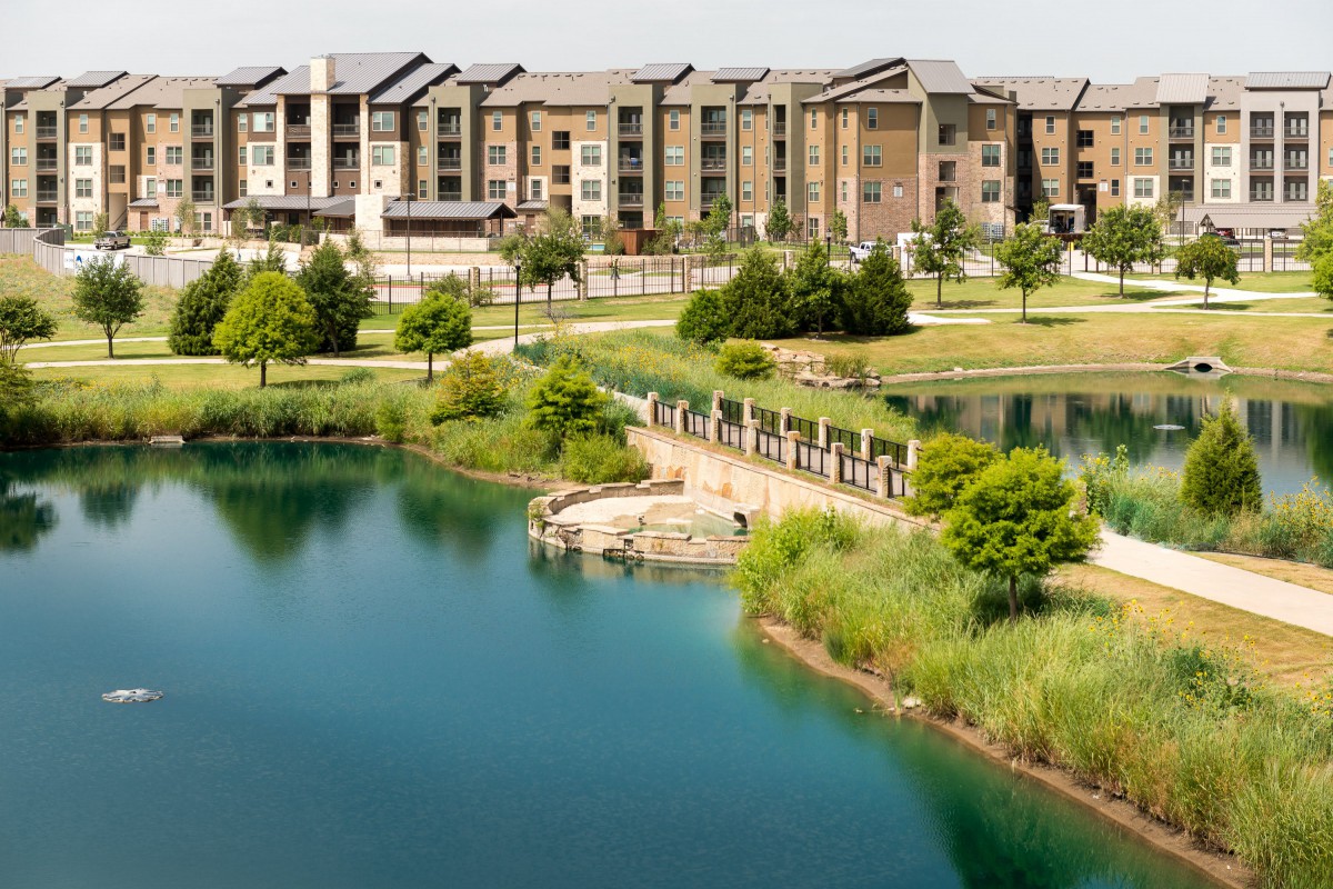 A scenic view of modern apartment buildings by a tranquil lake with a pedestrian bridge and lush greenery.
