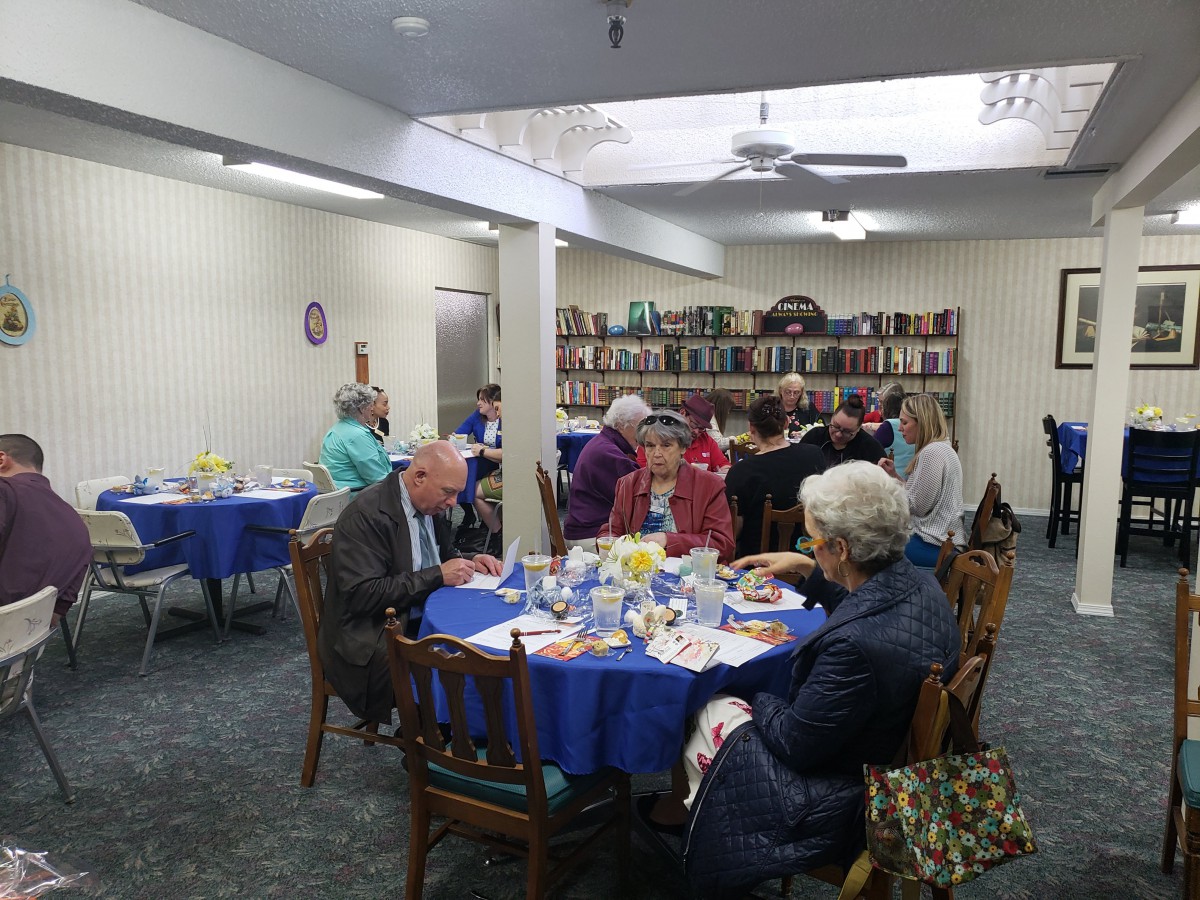 A group of people enjoying a meal together in a room with bookshelves and blue tablecloths.
