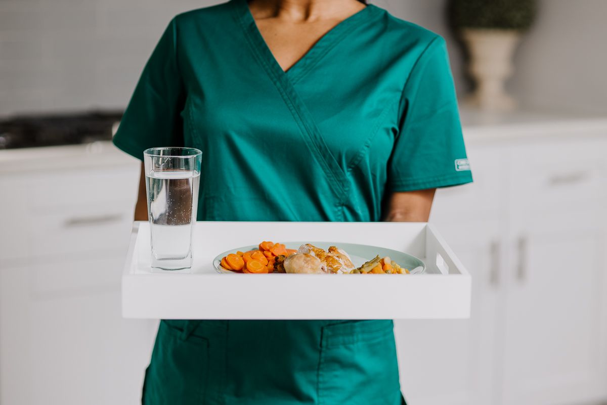 A healthcare worker in a green uniform carries a meal tray with water and healthy food.