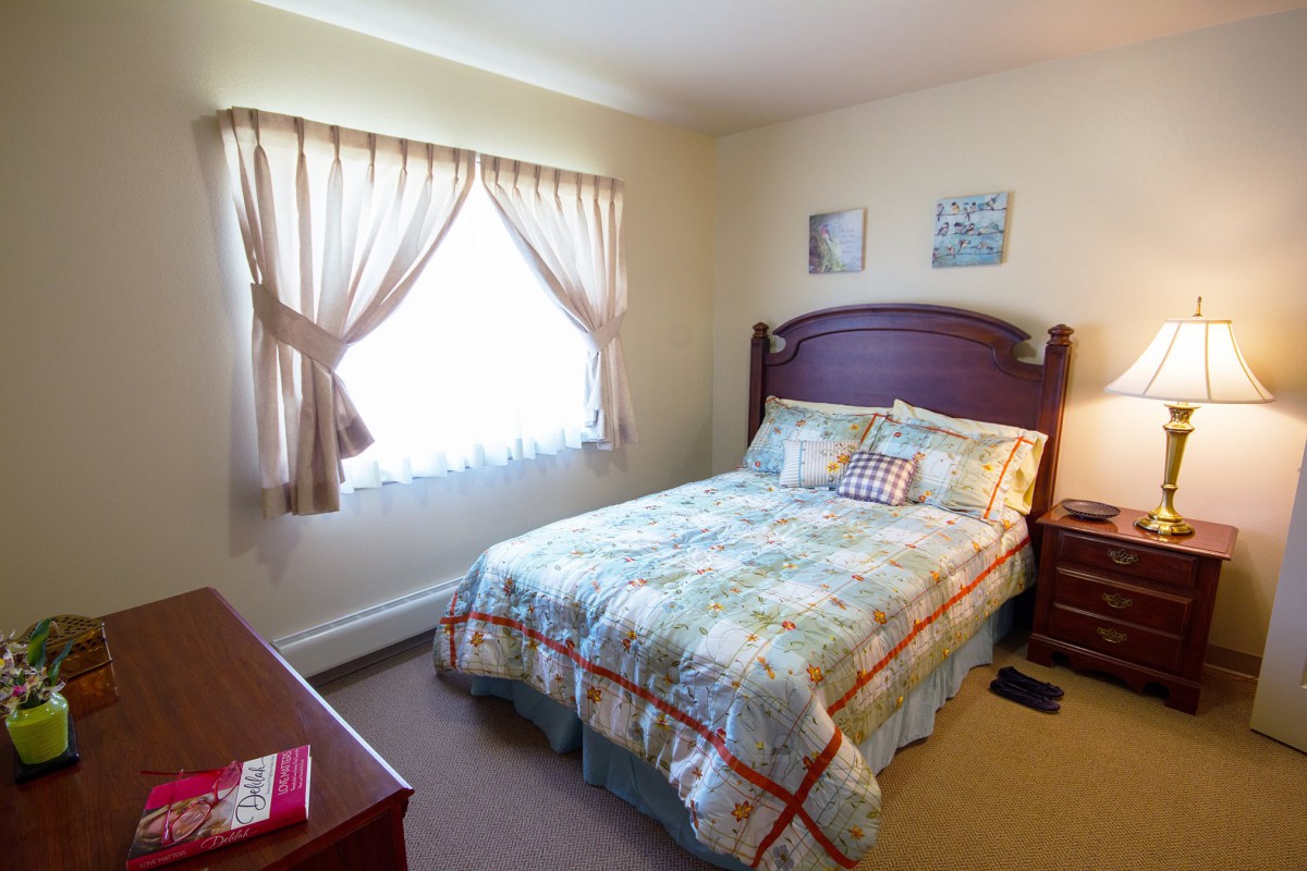 A cozy bedroom with a neatly made bed, soft lighting from a table lamp, and natural light streaming through the window with curtains.