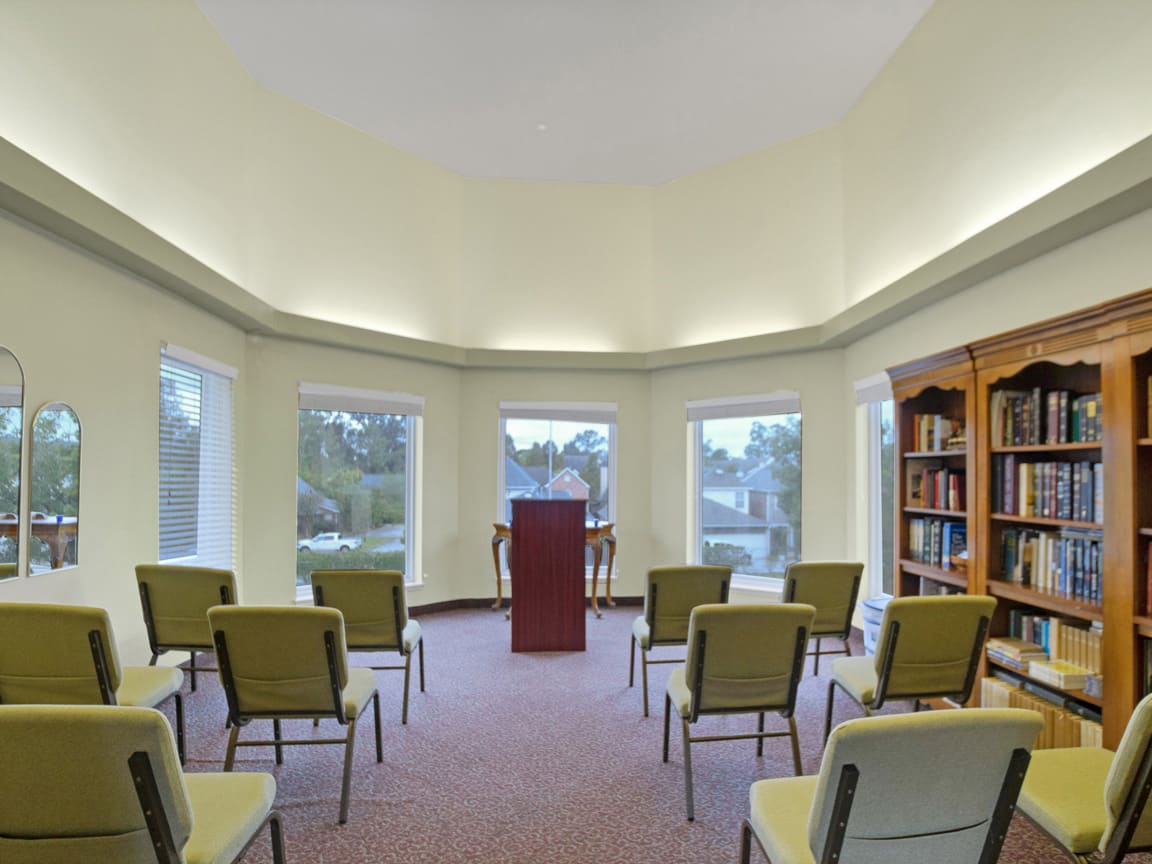 A small meeting room with a podium and several chairs arranged in rows, surrounded by windows and a bookshelf.