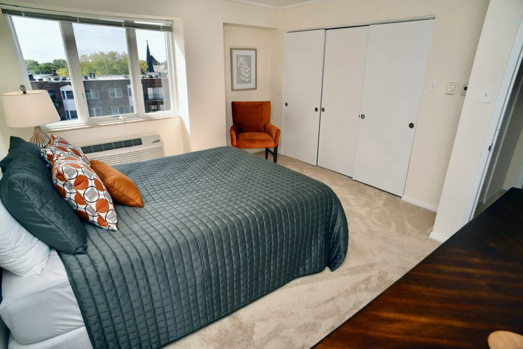 A cozy bedroom featuring a neatly made bed, an orange chair by the window, and a spacious closet with white doors.
