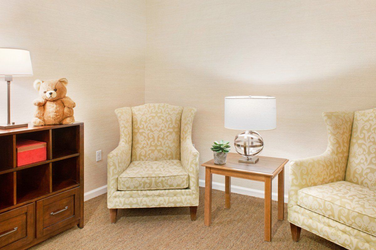 A cozy living room corner featuring a patterned armchair, a wooden side table with a lamp and plant, and nearby shelving.