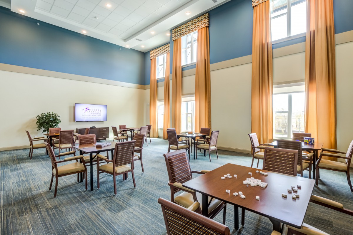 A spacious room with several tables and chairs, large windows with curtains, and a wall-mounted television.