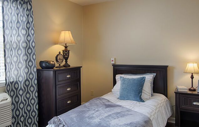 A cozy bedroom with a single bed, featuring a blue pillow and a dresser topped with a lamp.