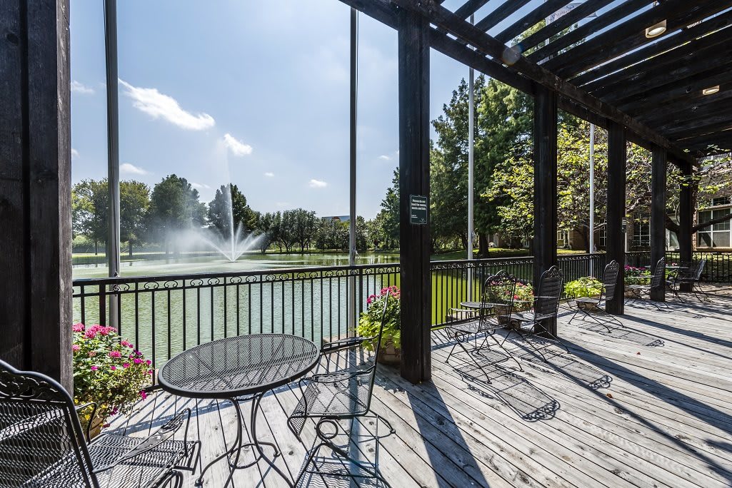 A scenic view of a fountain in a pond from an outdoor deck with seating, surrounded by trees and shaded by a pergola.