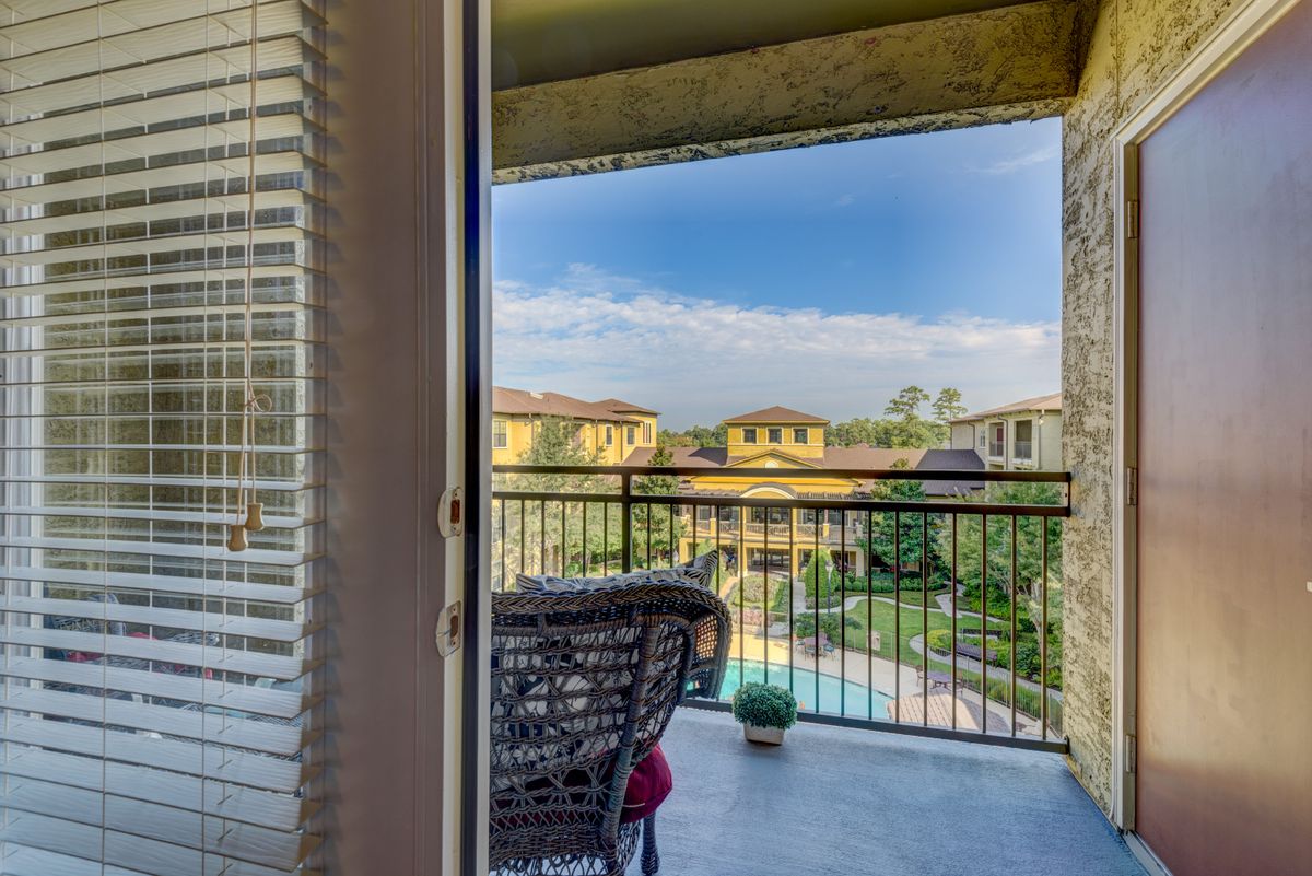 A cozy balcony with a rattan chair overlooks a residential area and pool under a blue sky.