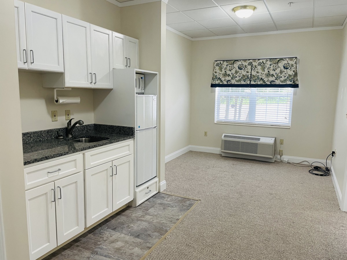 A small apartment interior featuring a kitchen with white cabinets, a sink, and a refrigerator. The room has carpet flooring and a window allowing natural light.