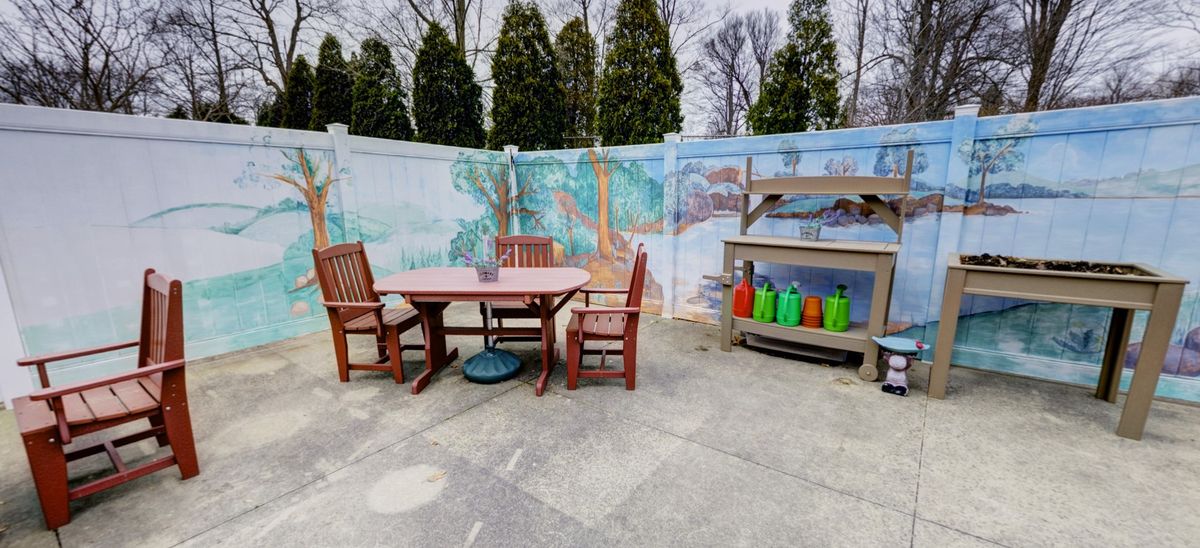 An outdoor patio with a wooden table and chairs, adjacent to a barbecue grill, set against a mural-painted fence.