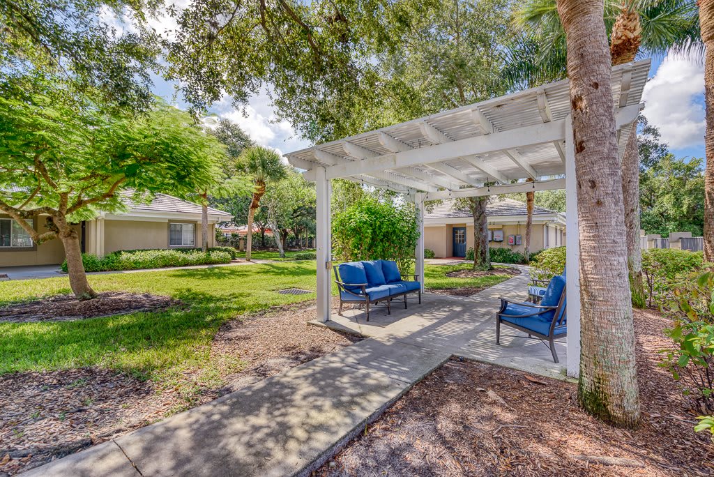 A serene garden features a white pergola with blue cushioned chairs, surrounded by lush greenery and trees.