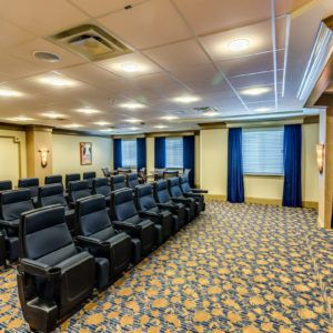 A well-lit conference room with multiple rows of comfortable seating and a carpeted floor.