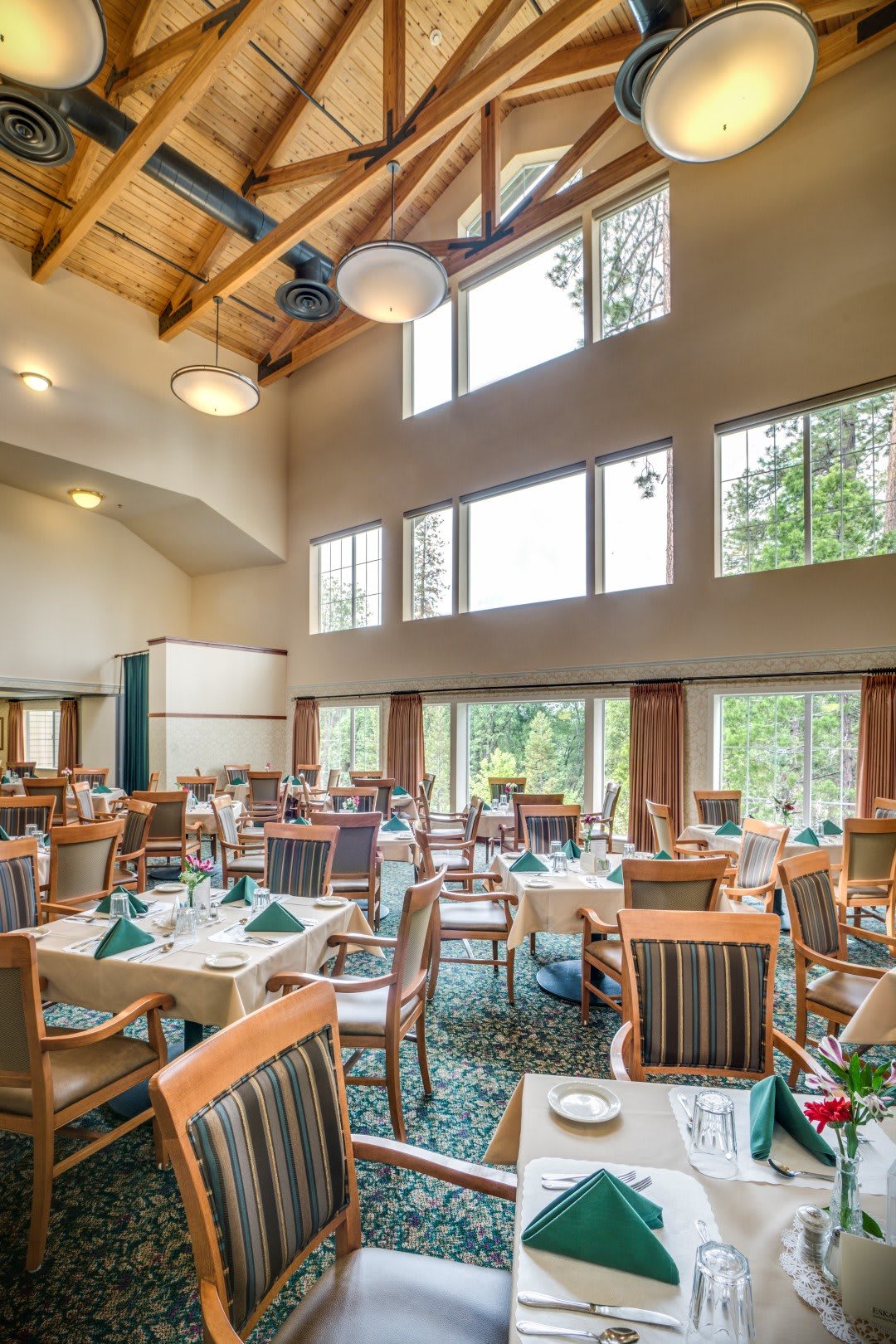 A spacious restaurant interior featuring a high wooden ceiling and large windows, with neatly arranged tables and chairs.