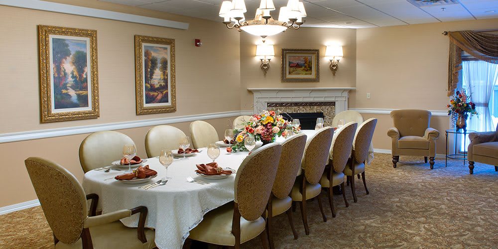 An elegantly set dining room with a long table adorned with floral arrangements, surrounded by upholstered chairs and illuminated by chandeliers.