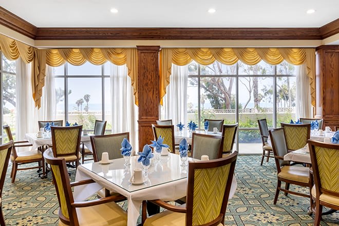 A beautifully arranged restaurant dining area with tables set for guests, featuring large windows and elegant curtains.