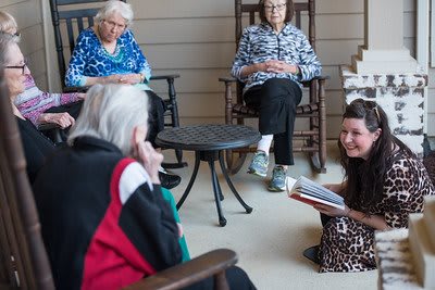 A group of elderly women sitting in rocking chairs, engaged in a reading session with a young woman.