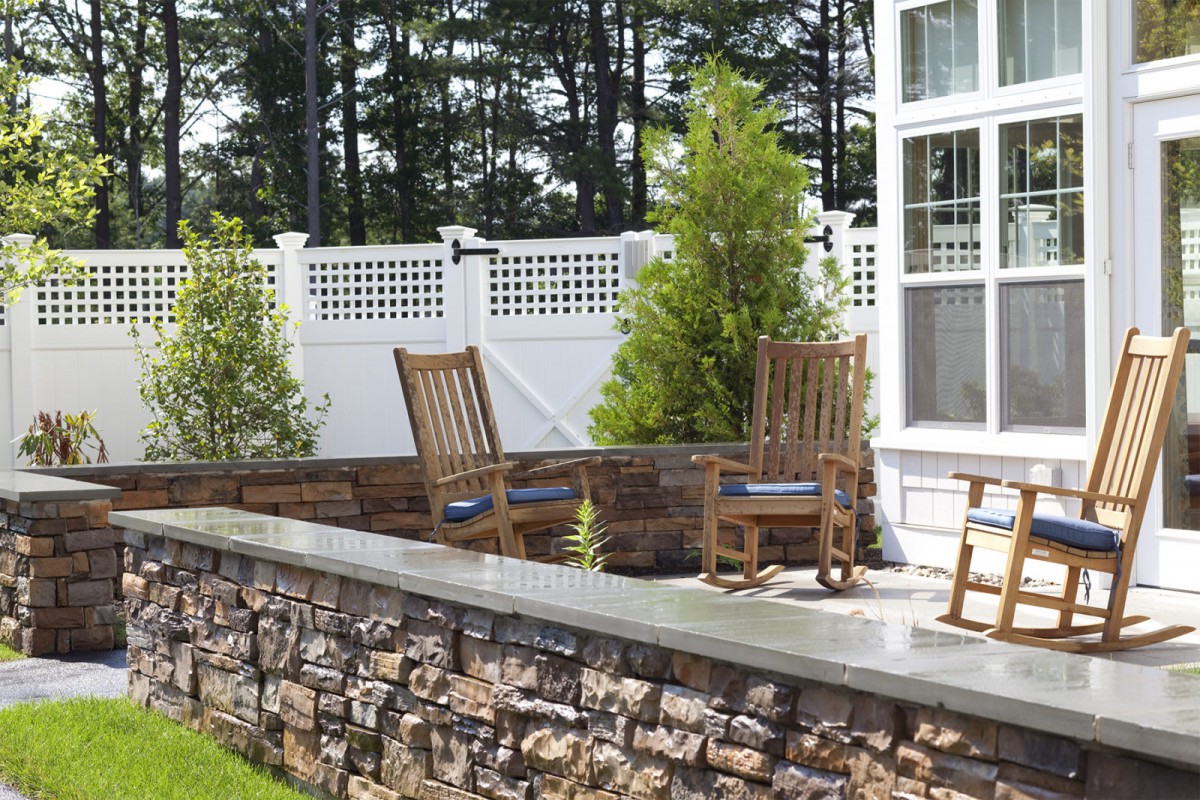 A serene outdoor patio area with wooden chairs, a stone wall, and greenery.