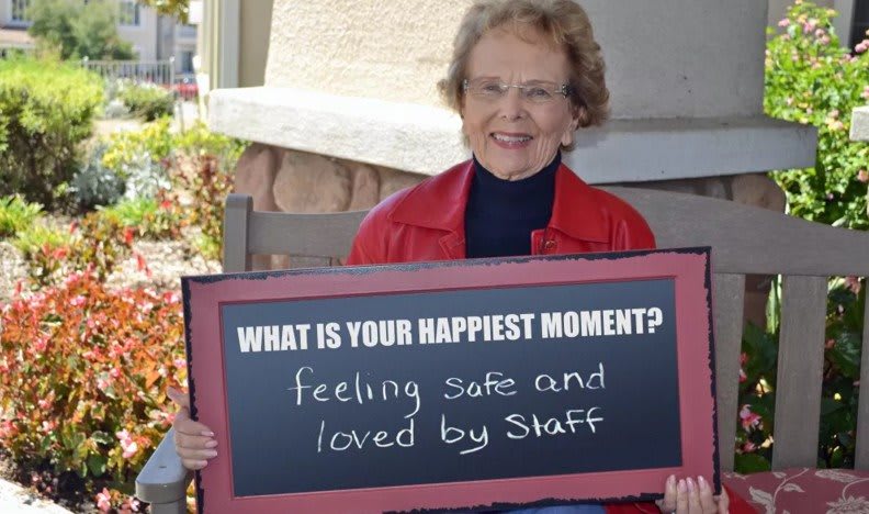 An elderly woman smiles as she holds a sign expressing her happiest moment of feeling safe and loved by staff.