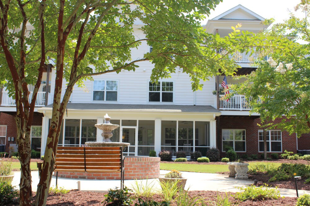 A peaceful outdoor area featuring a building facade, a fountain, and benches surrounded by trees and greenery.
