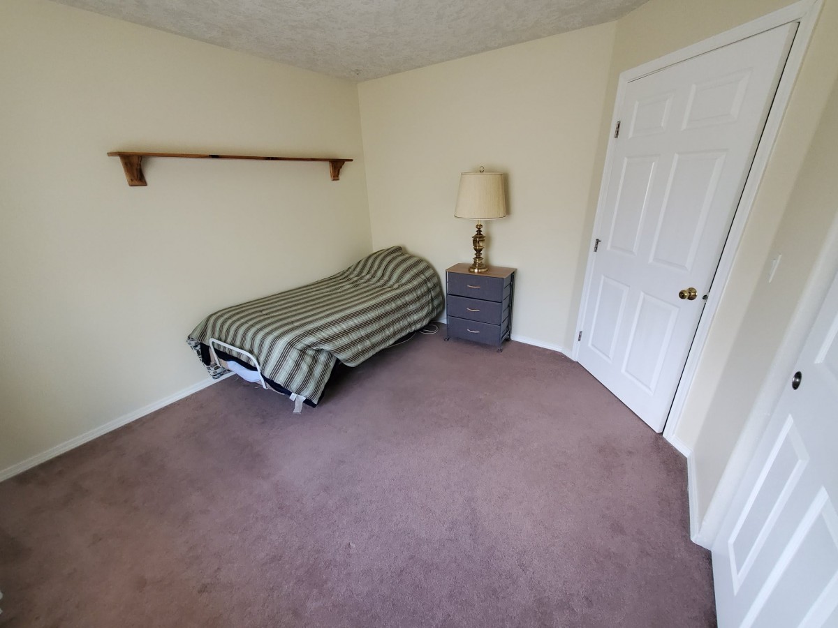 A minimalist bedroom with a single bed, carpeted floor, and a small bedside table with a lamp.
