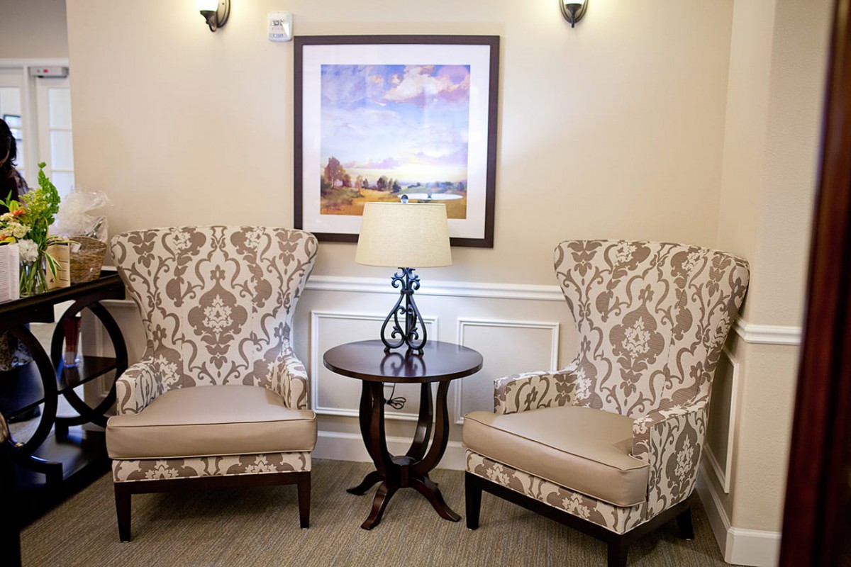 A cozy sitting area with two patterned chairs and a small round table featuring a decorative lamp, set against a wall with framed artwork.