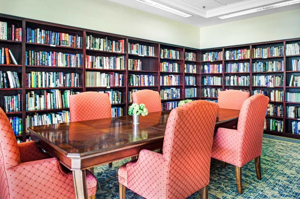 A cozy library reading room with shelves full of books and a wooden table surrounded by upholstered chairs.