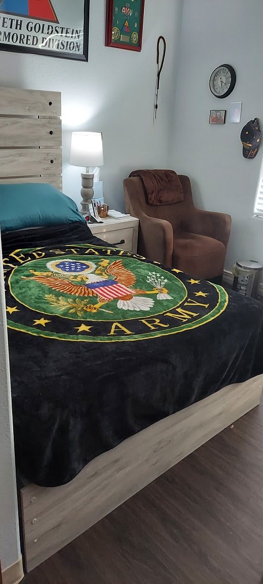 A bedroom featuring a US Army-themed blanket with an eagle emblem on the bed, alongside a brown chair and a nightstand.