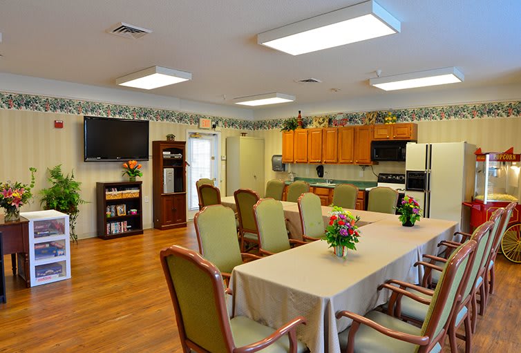 A spacious dining room with a table and chairs, adjacent to a kitchen area, decorated with flowers and featuring a television and bookshelf.