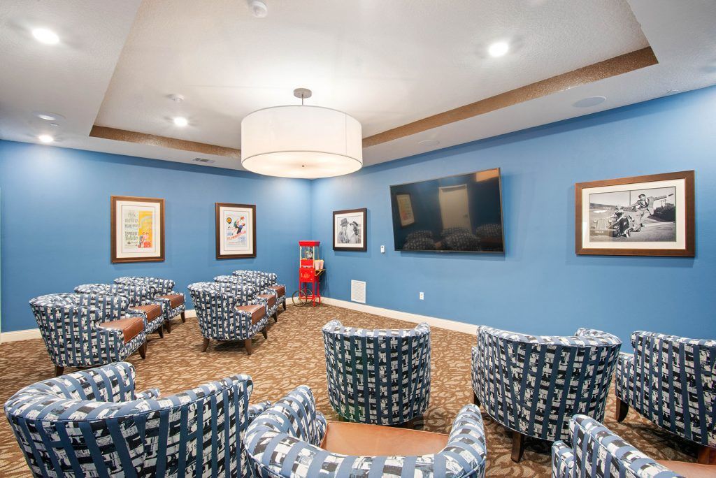 A cozy lounge area with patterned chairs arranged around a large television on blue-painted walls, adorned with framed artwork and illuminated by a central overhead light fixture.
