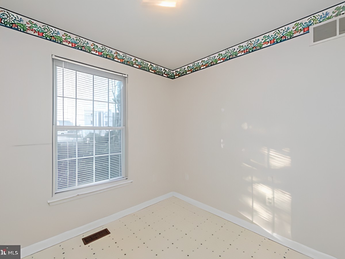 A small empty room with a window, wallpaper border, and floor vent.