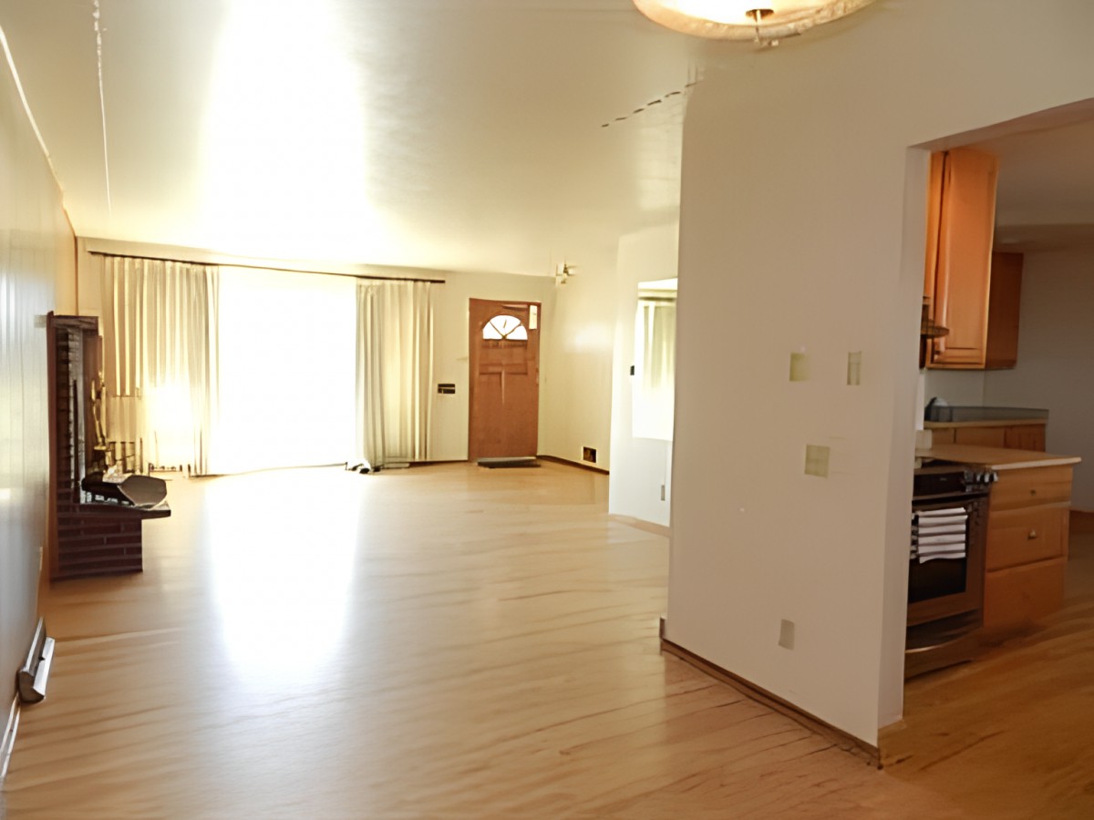 A bright living room with hardwood flooring, natural light streaming through large windows, and an entrance to the kitchen.