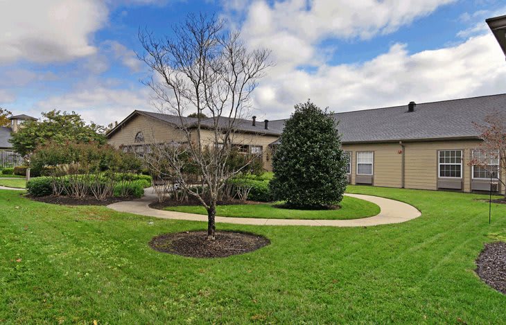 A residential house with a well-maintained lawn, a tree, and a curving walkway under a partly cloudy sky.