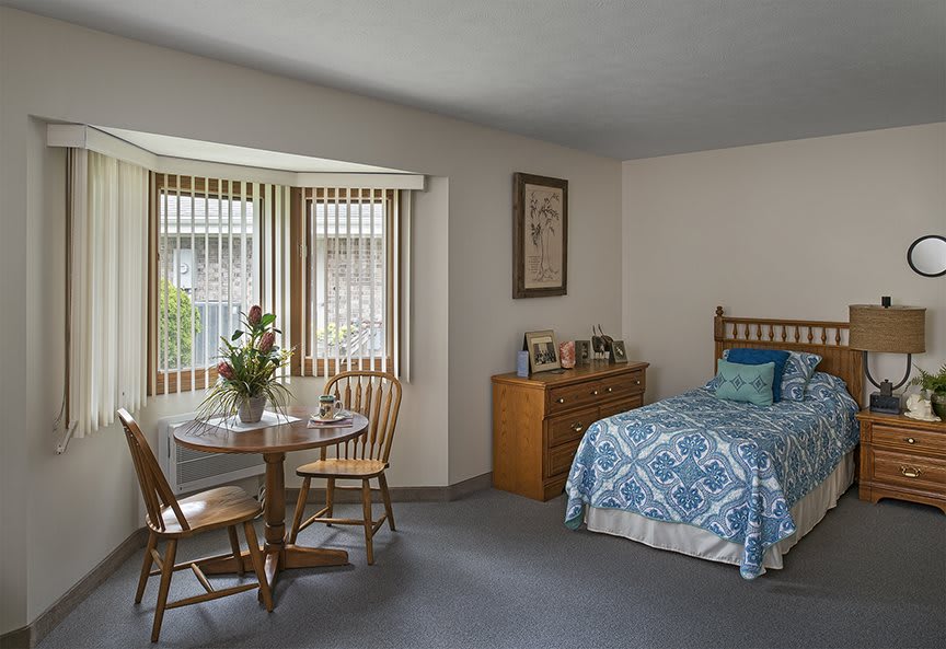 A cozy bedroom with a single bed, a small table with chairs by the window, and a dresser with framed photos.