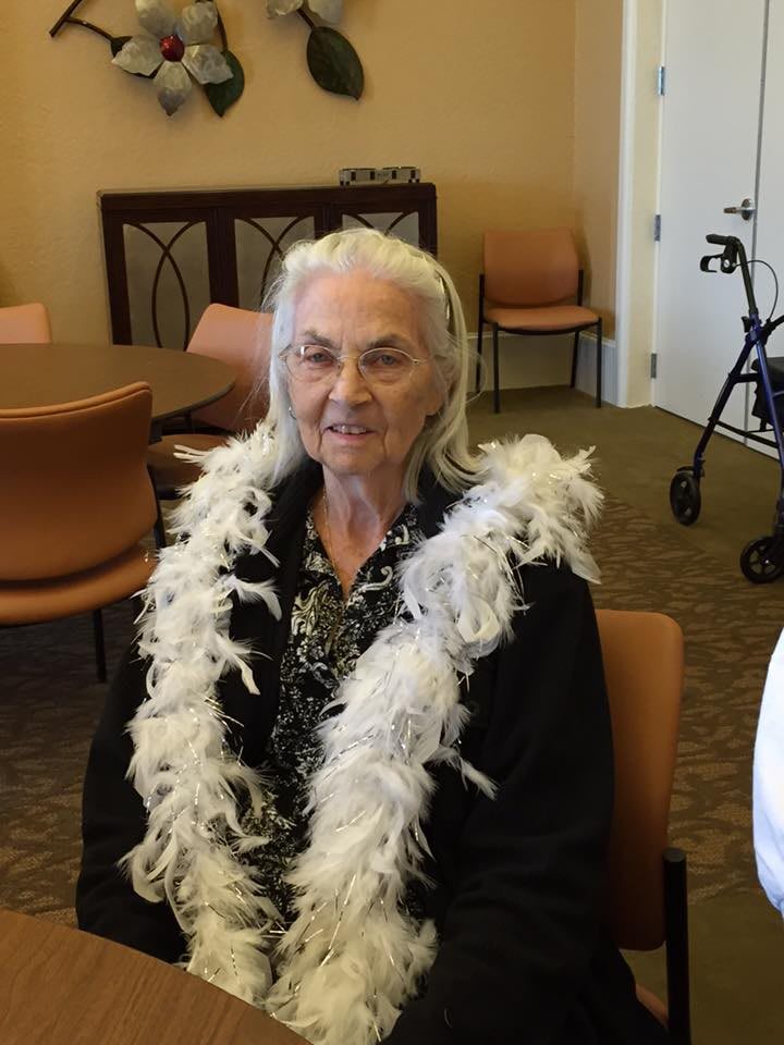 An elderly woman sitting indoors, wearing a feather boa and smiling.