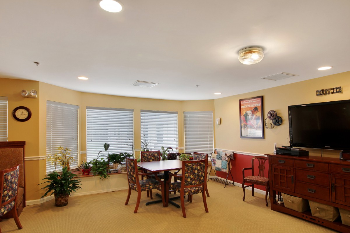 A cozy dining room with a table and chairs, surrounded by large windows and decorated with plants and artwork.