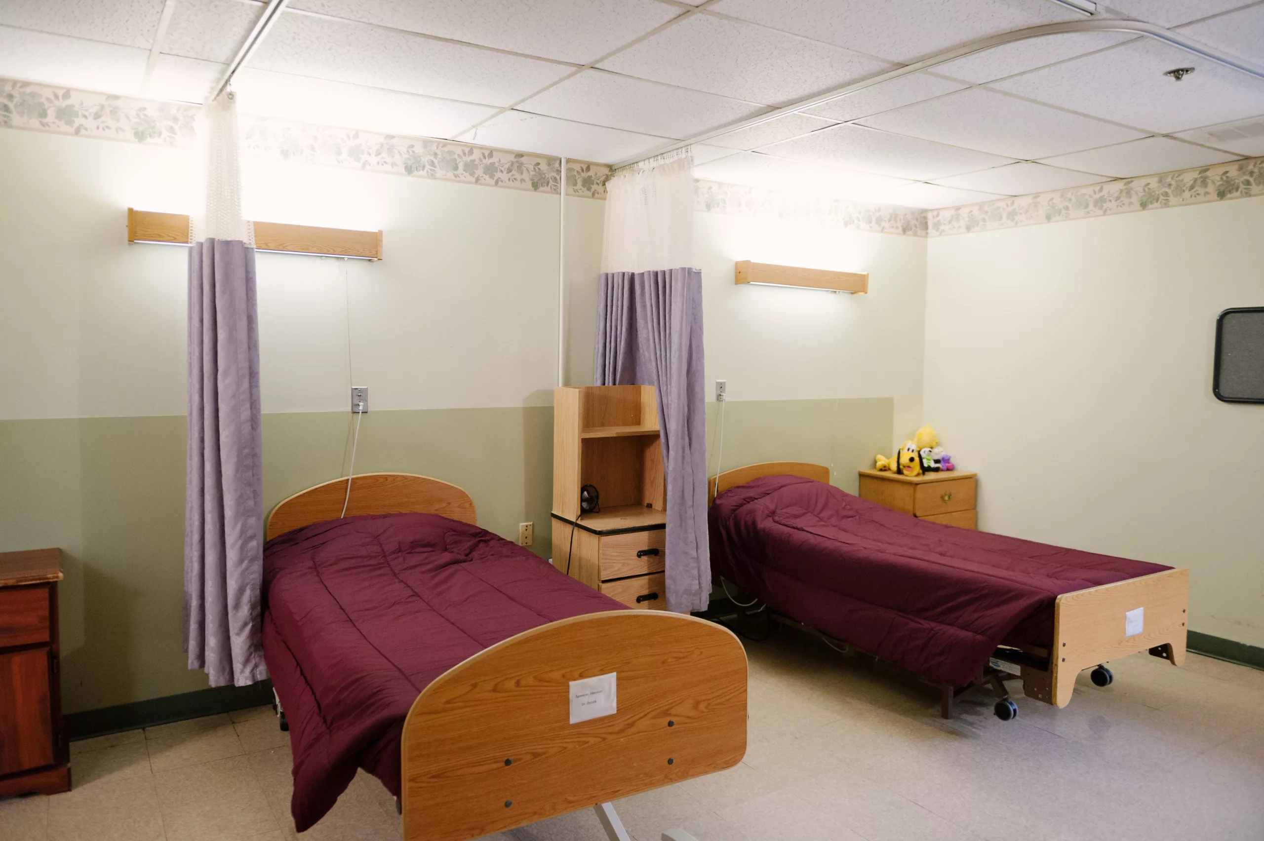 A hospital room with two twin beds, separated by a curtain, and simple wooden furniture.