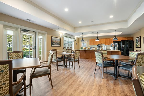 A spacious dining area with wooden flooring, featuring multiple tables and chairs, adjacent to a modern kitchen.