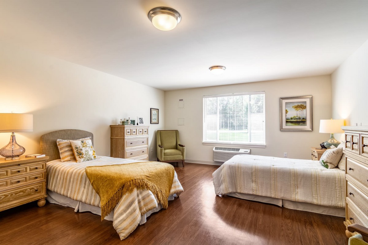A cozy bedroom with two single beds, a window providing natural light, a dresser, and an armchair.