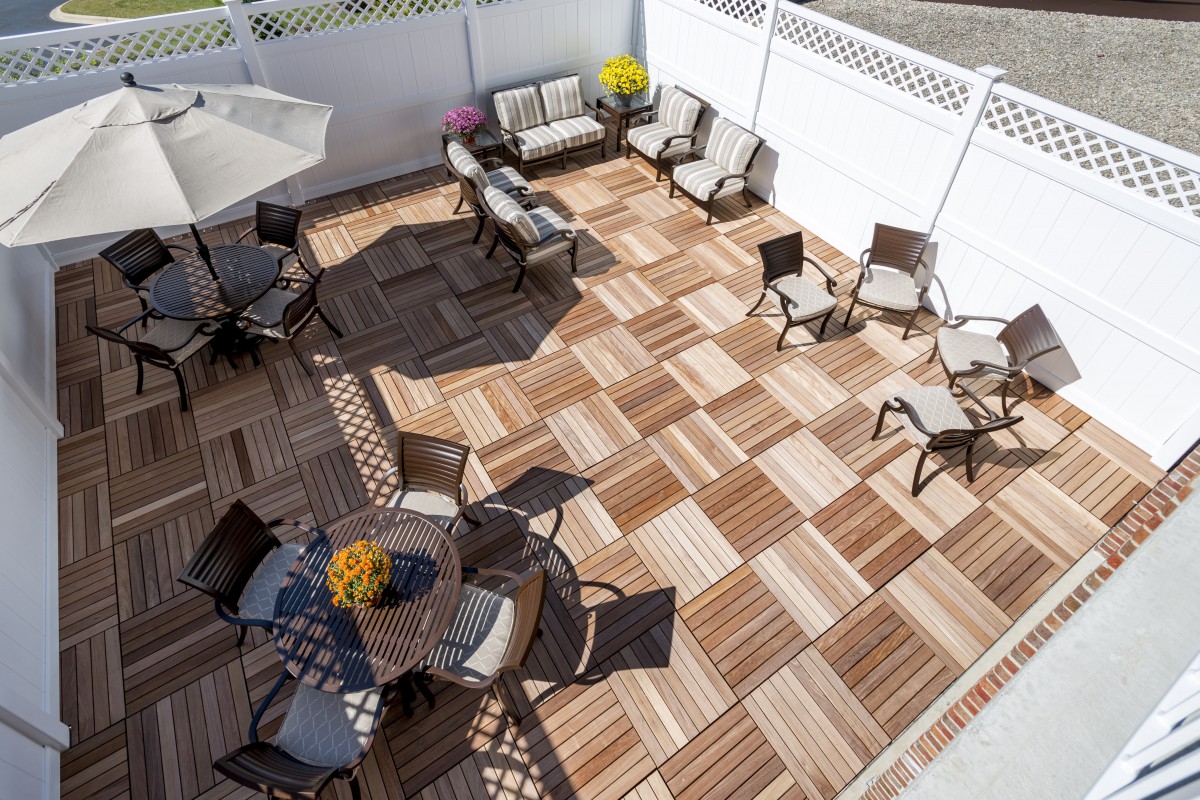 A spacious outdoor patio with wooden deck tiles, featuring various seating arrangements including chairs, a table with an umbrella, and a comfortable sofa set against a white fence.
