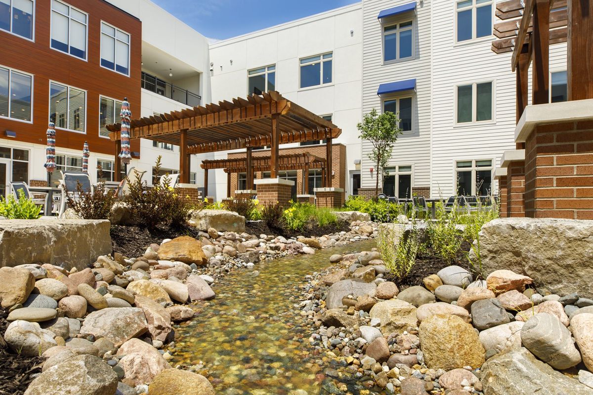 A modern apartment complex features a serene outdoor garden with a pergola and a small stream flowing through rocks.
