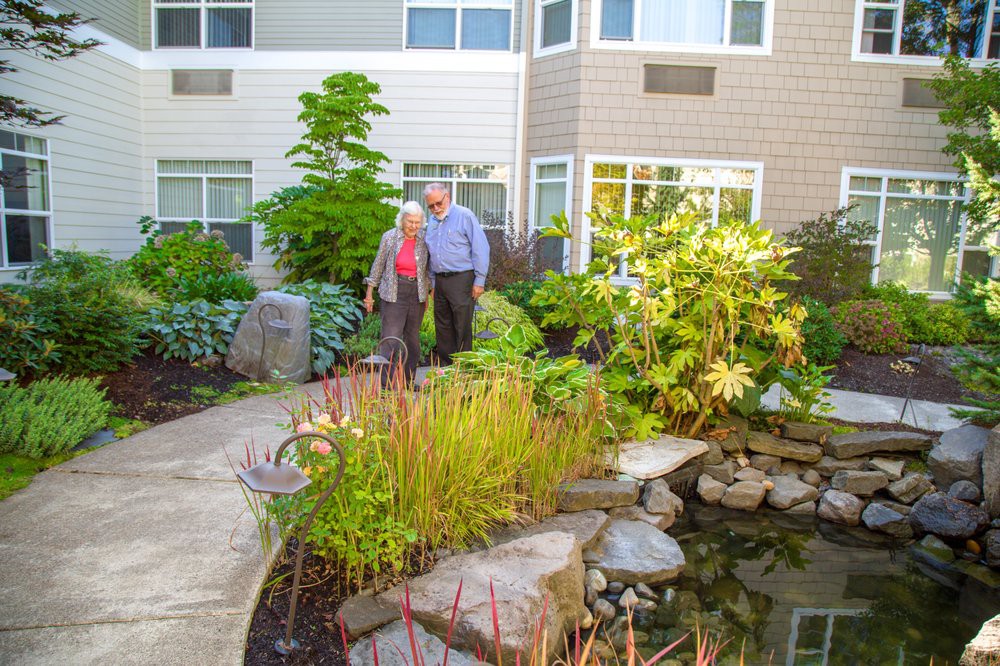 An elderly couple enjoys a walk through a lush garden with a pond in a residential area.