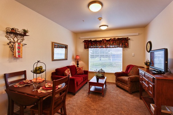 A cozy living room with a sofa, armchair, and dining area, featuring warm tones and a large window overlooking greenery.