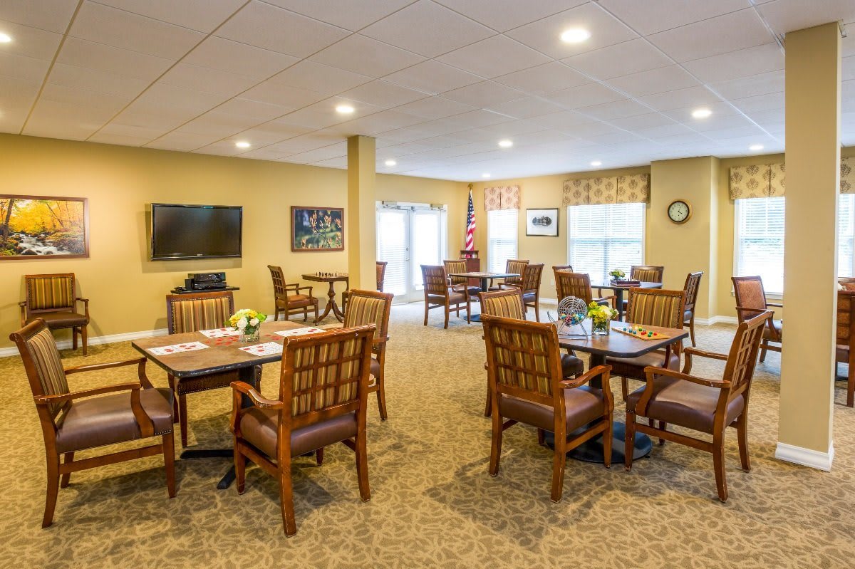 A cozy dining room with wooden tables and chairs, featuring bright lighting and a television on the wall.