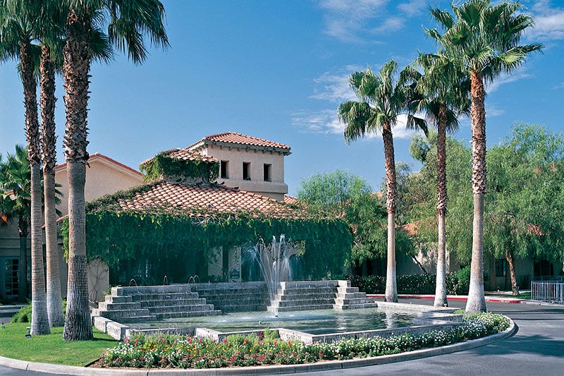 A picturesque building with a vine-covered facade, accompanied by a cascading fountain and palm trees under a clear blue sky.