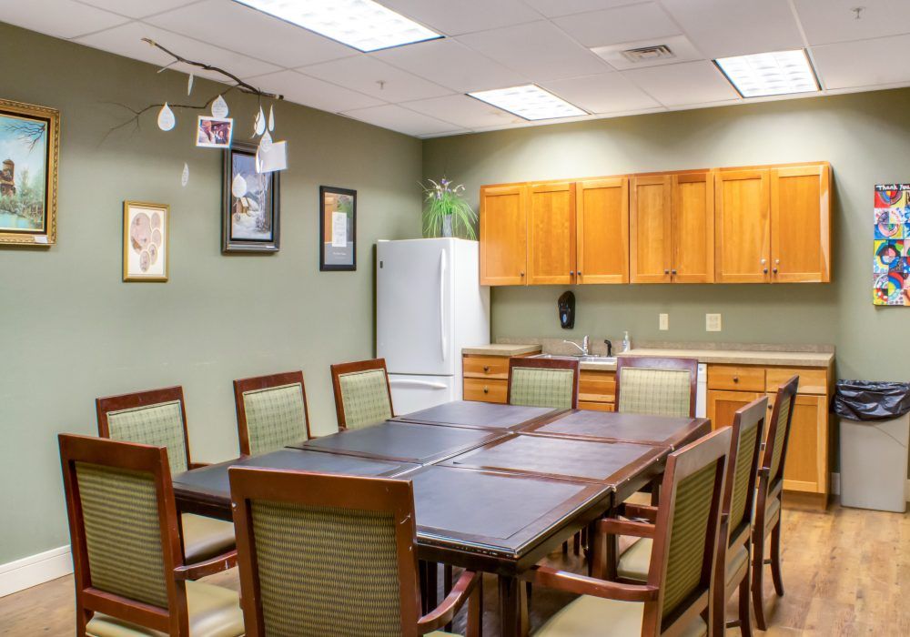 A conference room with a large table and chairs, adjacent to a kitchenette featuring cabinets and a refrigerator.