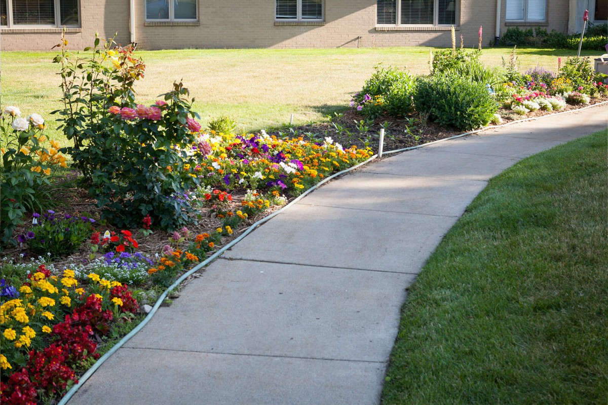 A curved sidewalk bordered by a colorful flower garden in a residential area.
