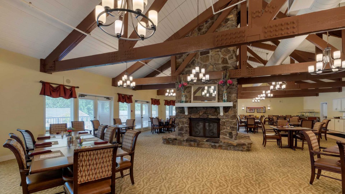 A spacious dining and lounge area with a central fireplace, high ceilings, chandeliers, and wooden beams.